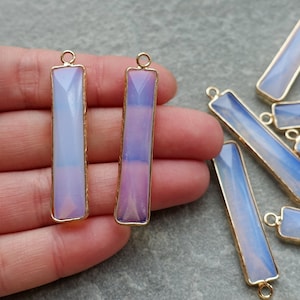 Puede incluir: Colgantes rectangulares de piedras preciosas opalescentes facetadas con bordes de metal dorado. Las piedras preciosas hacen la transición de azul claro a púrpura. Cada colgante tiene un pequeño lazo en la parte superior para ensartar. Varios colgantes son visibles.