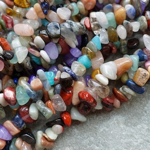 May include: A close-up shot of a selection of gemstone chip beads in a variety of colours. The beads are in different shapes and sizes, including red, blue, purple, white, and orange. They are strung together, possibly for jewellery.
