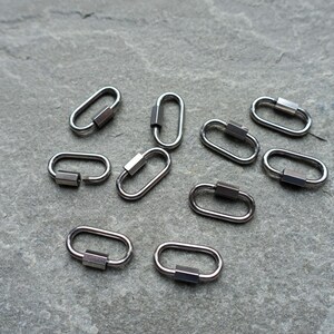K&ouml;nnte beinhalten: Eine Ansammlung von neun kleinen, ovalen Metall-Karabinerverschl&uuml;ssen in einem dunklen Gunmetal-Grau. Jeder Verschluss verf&uuml;gt &uuml;ber einen federbelasteten Schlie&szlig;mechanismus an einer Seite, der zum sicheren Verbinden von Schmuckteilen dient.