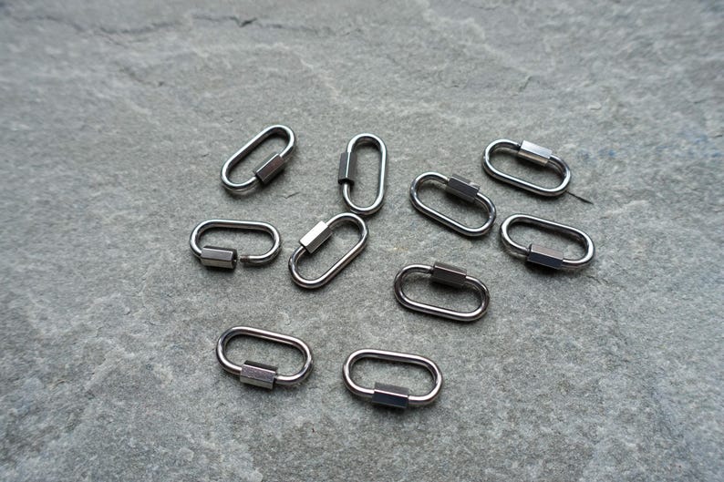 K&ouml;nnte beinhalten: Eine Ansammlung von neun kleinen, ovalen Metall-Karabinerverschl&uuml;ssen in einem dunklen Gunmetal-Grau. Jeder Verschluss verf&uuml;gt &uuml;ber einen federbelasteten Schlie&szlig;mechanismus an einer Seite, der zum sicheren Verbinden von Schmuckteilen dient.