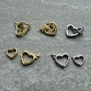 Könnte beinhalten: Eine Auswahl an herzförmigen Schmuckverschlüssen in Gold- und Silberfarben. Die Verschlüsse sind in verschiedenen Größen und Stilen erhältlich, darunter ineinandergreifende und einteilige Designs. Präsentiert auf einer strukturierten, grauen Oberfläche.