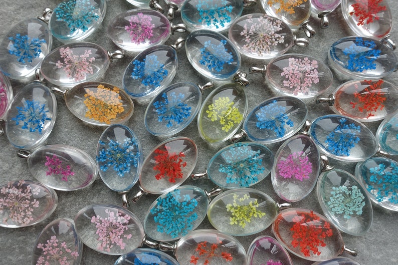 Op de afbeelding: Een verzameling ovale hangers van heldere hars, elk met gedroogde bloemen in verschillende kleuren, waaronder roze, blauw, geel en rood. Elke hanger heeft een zilverkleurige oogje. De hangers liggen op een grijze ondergrond.