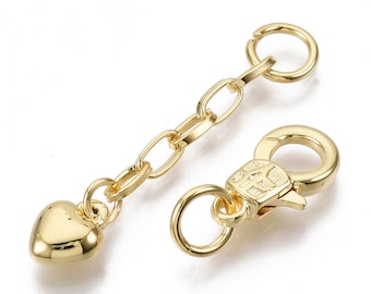 May include: A gold-colored jewelry clasp with a heart-shaped charm and chain. The clasp has a lobster claw closure and a small ring. The chain is made of oval links. The heart charm is smooth and rounded.