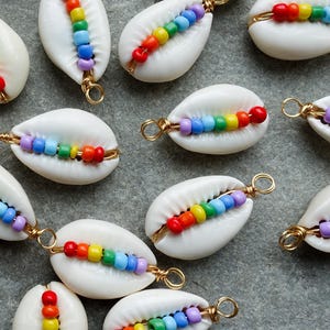 May include: White cowrie shells decorated with a rainbow of colourful beads. Each shell has a gold-coloured wire loop for hanging. The beads are arranged in a linear pattern, creating a vibrant design. Ideal for jewellery making or crafting projects.