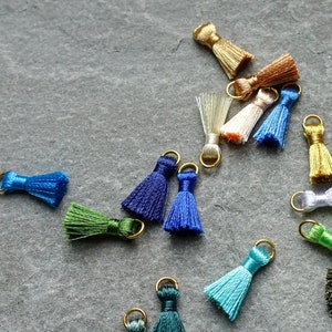 May include: A collection of small, colorful tassels with gold-colored metal loops. The tassels are in various shades, including blue, green, gold, and white. They are scattered on a gray, textured surface, suitable for jewelry making or craft projects.