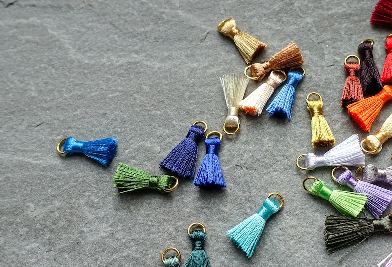 May include: A collection of small, colorful tassels with gold-colored metal loops. The tassels are in various shades, including blue, green, gold, and white. They are scattered on a gray, textured surface, suitable for jewelry making or craft projects.