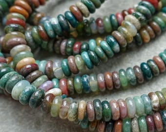 May include: Close-up of several strands of gemstone beads. The beads are a mix of colors, including green, red, brown, and cream. The beads are small and round, and appear to be strung together. The background is a light gray surface.