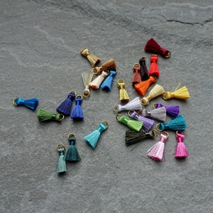 May include: A collection of small, colorful tassels with gold-colored metal loops. The tassels are in various shades, including blue, green, pink, purple, and yellow. They are scattered on a gray, textured surface.