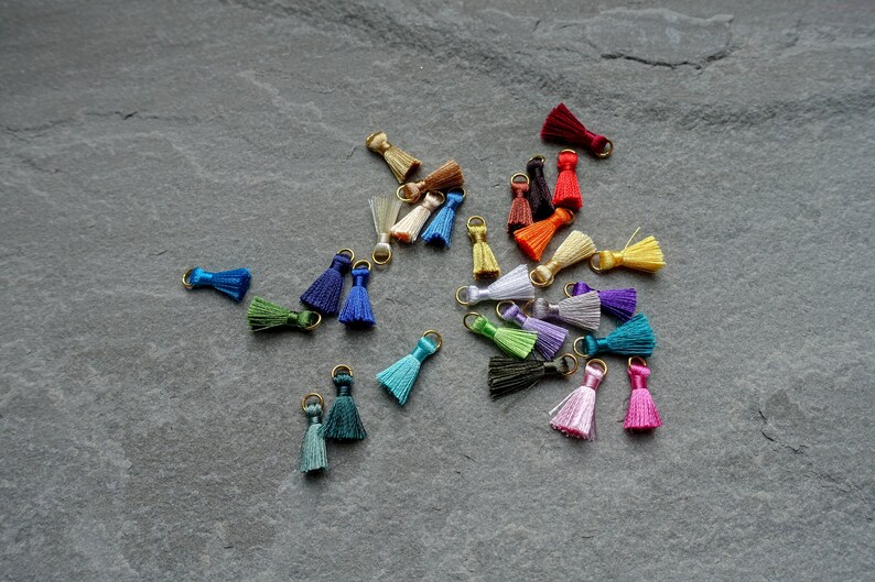 May include: A collection of small, colorful tassels with gold-colored metal loops. The tassels are in various shades, including blue, green, pink, purple, and yellow. They are scattered on a gray, textured surface.