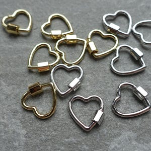 May include: An assortment of heart-shaped carabiner clasps in gold and silver tones. The clasps are open and ready to be attached to a necklace or bracelet. The clasps are made of metal and are on a gray surface.