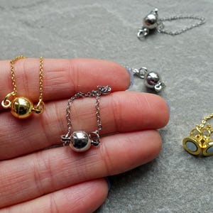 Könnte beinhalten: Nahaufnahme mehrerer kleiner Magnetverschlüsse für Schmuck. Die Verschlüsse sind in Gold- und Silbertönen gehalten und haben ein kugelförmiges Design. Die Verschlüsse sind durch kleine Ketten verbunden. Der Hintergrund ist eine graue Oberfläche.