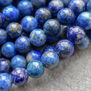 May include: A close-up of a collection of round lapis lazuli beads. The beads are a deep blue colour with flecks of gold and white, and are approximately 8mm in diameter. The beads are strung together, possibly for jewellery making.
