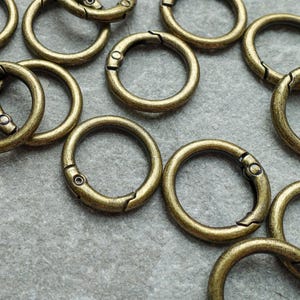 Op de afbeelding: Een verzameling antiek messingkleurige ronde veersnapsluitringen. Deze metalen ringen hebben een scharnierende opening en een veergeladen sluiting. De ringen zijn verspreid over een grijze ondergrond.