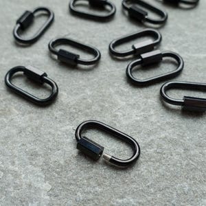 K&ouml;nnte beinhalten: Eine Ansammlung kleiner, ovaler Schraubkarabiner aus schwarzem Metall. Jeder Karabiner verf&uuml;gt &uuml;ber eine glatte, gl&auml;nzende Oberfl&auml;che und einen Gewindemechanismus, durch den sich die Schlaufe sicher &ouml;ffnen und schlie&szlig;en l&auml;sst.