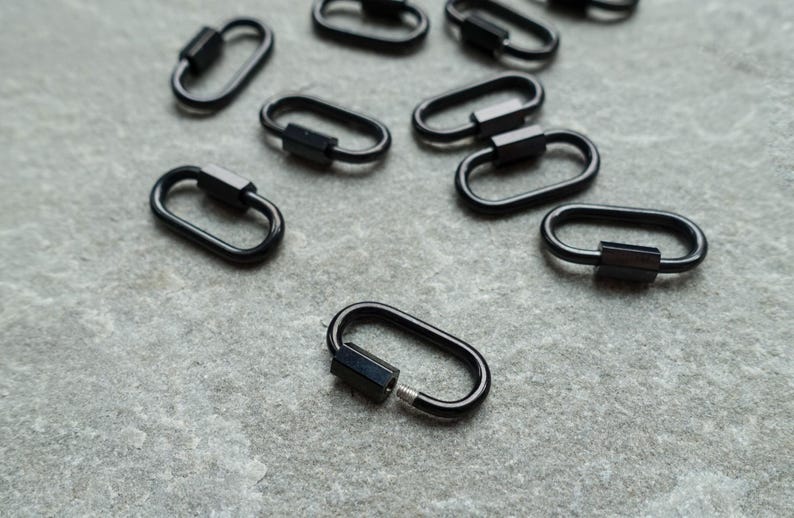K&ouml;nnte beinhalten: Eine Ansammlung kleiner, ovaler Schraubkarabiner aus schwarzem Metall. Jeder Karabiner verf&uuml;gt &uuml;ber eine glatte, gl&auml;nzende Oberfl&auml;che und einen Gewindemechanismus, durch den sich die Schlaufe sicher &ouml;ffnen und schlie&szlig;en l&auml;sst.