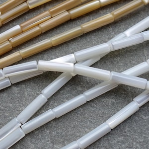 以下が含まれることがあります： 白とタンの色合いの長方形の、虹色のビーズのストランドのクローズアップ。ビーズは一緒にひもでつながれており、質感のある表面を作り出しています。ビーズは微妙な光沢があり、光を反射しています。