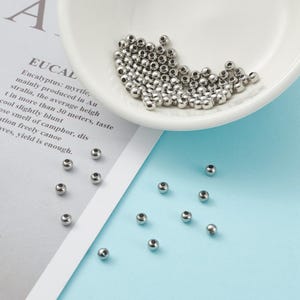 Peut inclure: Un ensemble de petites perles m&eacute;talliques brillantes de couleur argent&eacute;e. Certaines perles sont dans un plat blanc incurv&eacute;, tandis que d'autres sont dispers&eacute;es sur une surface bleu clair. Les perles ont une forme lisse et arrondie.