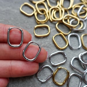 Op de afbeelding: Zilveren en goudkleurige ovale oorringen. De oorbellen worden getoond op een grijze ondergrond, met een hand die twee zilveren oorbellen vasthoudt. Extra oorbellen in goud en zilver zijn verspreid.