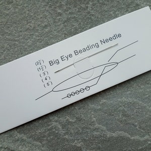 May include: A white card with the text "Big Eye Beading Needle" and a diagram of a needle being threaded. The card also lists needle sizes in inches. A single silver needle is visible.