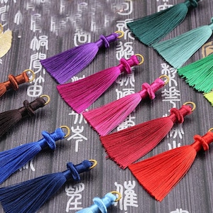 May include: A collection of colorful tassels in various shades, including purple, pink, red, blue, and green. Each tassel features a looped top with a gold-colored ring. The tassels are arranged on a dark surface with Asian-style characters.