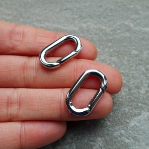 Op de afbeelding: Twee glanzende, zilverkleurige ovale karabijnhaakclips. De clips zijn gemaakt van metaal en hebben een glad, reflecterend oppervlak. Ze worden getoond tegen een neutrale achtergrond.