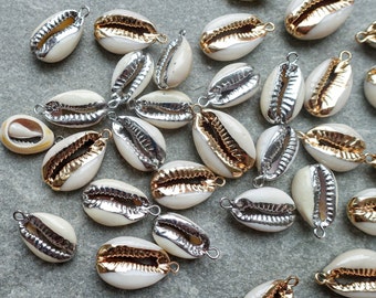 May include: A collection of small, white cowrie shell pendants with metallic gold and silver edges. Each shell has a loop for stringing. The shells are scattered on a gray surface, showcasing their natural texture and contrasting metallic accents.