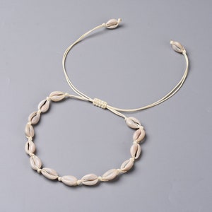 May include: A cream-coloured cowrie shell necklace and choker set. The necklace features multiple shells strung together, while the choker has a single shell at each end. The set is connected by a cream-coloured cord.