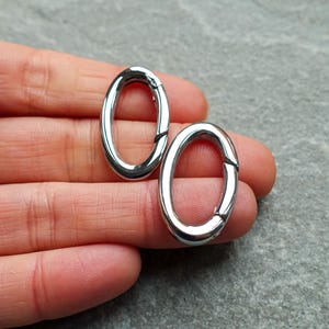 May include: Two silver-toned oval carabiner rings with a polished, reflective finish. Each oval ring features a spring-loaded gate mechanism that allows the loop to open and close, providing a secure connection point for various hardware applications.