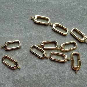 May include: A collection of gold-colored rectangular clasp connectors, scattered on a gray surface. Each clasp has a hinged design with a small loop at each end. These jewelry findings are ideal for necklaces and bracelets.