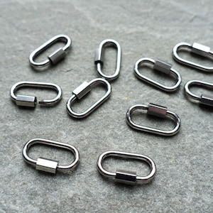 Puede incluir: Un conjunto de cierres de rosca ovalados de color plateado. Cada cierre presenta un mecanismo de rosca rectangular para un cierre seguro. Los cierres están dispuestos sobre una superficie gris texturizada, adecuados para la fabricación de joyas o manualidades.