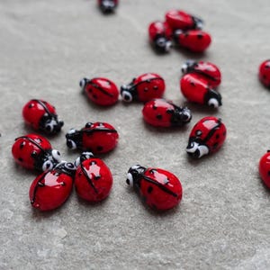 May include: A collection of small, red ladybug beads with black spots and black and white heads. The ladybugs are scattered on a gray surface, showcasing their glossy, detailed design. These decorative beads are ideal for crafting.