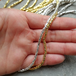 Peut inclure: Gros plan sur des brins de perles argentées et dorées. Les brins sont constitués de petites perles rectangulaires, tenues dans une main. Les perles ont un éclat métallique. Le fond est gris neutre.