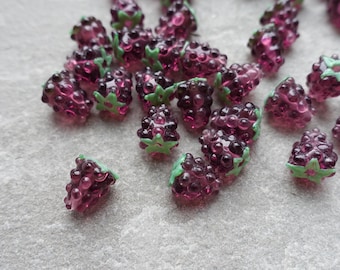 May include: A collection of translucent, purple grape-shaped beads with green leaf accents. The beads are clustered together, suggesting a jewelry-making theme. The beads are scattered on a light gray surface.