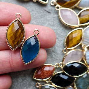 Puede incluir: Colgantes de piedras preciosas en forma de marquesa en varios colores, incluyendo ojo de tigre, azul, rosa y negro, engastados en biseles dorados. Estos colgantes están diseñados para la fabricación de joyas.