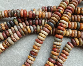 May include: Close-up of several strands of colorful, round gemstone beads. The beads are a mix of earth tones, including red, brown, tan, and gray. The beads are strung together, creating a textured and organic look.
