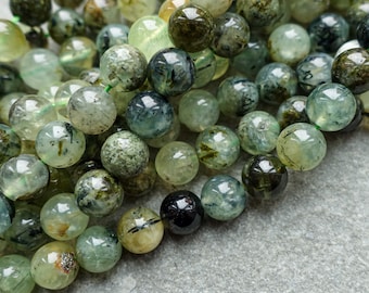 May include: Close-up of a strand of Prehnite gemstone beads. The beads are round, with a translucent, pale green color and dark inclusions. The beads are strung together with a green string, creating a natural and earthy aesthetic.