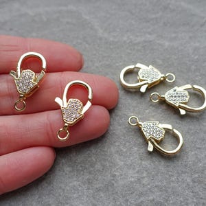 May include: Gold-tone lobster claw clasps featuring a Hamsa hand design, embellished with small, clear crystals. These jewellery findings are suitable for necklaces, bracelets, and other handmade projects. The clasps are shown on a grey surface.