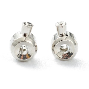 May include: Two shiny, silver-coloured metal cord end caps. Each cap has a cylindrical body with a central hole and a smaller, angled tube extending from the top. The caps are designed for securing cords or drawstrings.