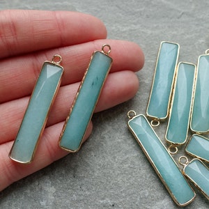 以下が含まれることがあります： 長方形の、薄い青色の宝石ペンダント。金色の金属フレーム付き。ペンダントはファセット加工された表面を持ち、ジュエリーに取り付けるための小さなループが上部にあります。宝石は柔らかく、くすんだ青緑色です。