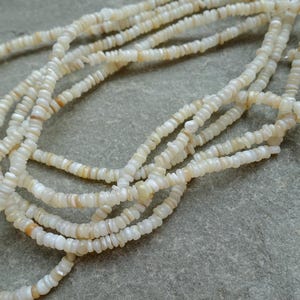 May include: Several strands of off-white, small, irregular beads. The beads are strung together, forming multiple necklaces or strands. The beads have a slightly iridescent quality, with hints of tan and cream colours. The beads are likely made of shell or a similar material.