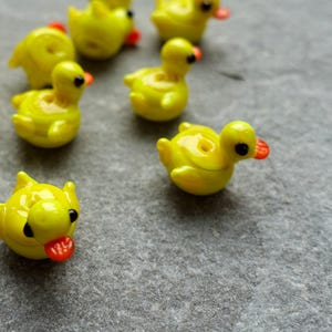 May include: Several small, yellow glass duck figurines with black eyes and orange beaks are scattered on a grey surface. Some ducks have their tongues out. The figurines are detailed and appear to be handmade, possibly for crafting or collecting.