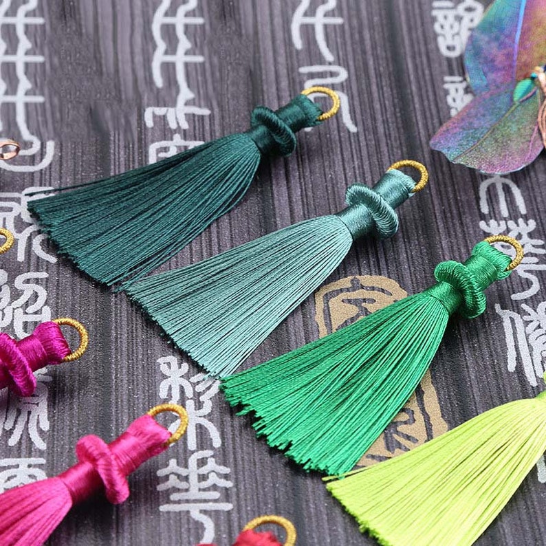 May include: Close-up of colorful silk tassels in shades of green, pink, and yellow. Each tassel has a gold-colored ring at the top. The background is a dark surface with Asian-style characters.