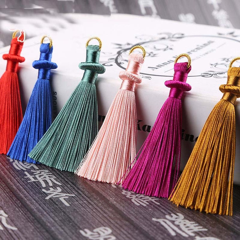 May include: A collection of colorful silk tassels in red, blue, green, pink, magenta, and gold. Each tassel has a decorative top with a gold-colored ring for hanging. The tassels are arranged in a row, showcasing their vibrant colors and textures.
