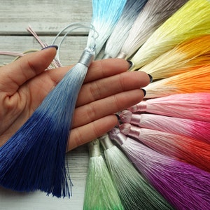 May include: A selection of vibrant silk tassels in a range of colours, including blue, pink, orange, and green. One tassel is held in a hand, displaying a blue ombre effect. The tassels have a silver metallic cap and are made of silky threads.