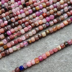 May include: A collection of small, square-cut gemstone beads in various shades of pink, purple, and brown. The beads are strung together, forming a textured surface. These beads are likely used for jewellery making.