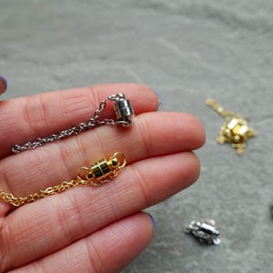 May include: Close-up of several magnetic jewellery clasps in silver and gold tones. The clasps are attached to delicate chain necklaces. The clasps are cylindrical and have a magnetic closure. The background is a neutral grey.