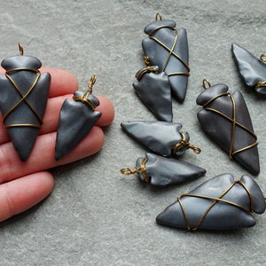 May include: Handmade grey arrowhead pendants with gold wire wrapping. The pendants have a metallic sheen and are arranged on a grey surface. These unique pendants are ideal for jewellery making or as decorative accents.