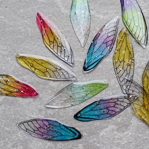 Puede incluir: Una colección de alas de hadas coloridas y translúcidas con intrincados detalles de venas negras y acentos de purpurina dorada. Las alas presentan un degradado de colores, incluyendo rojo, amarillo, azul y verde, creando una estética caprichosa y etérea.