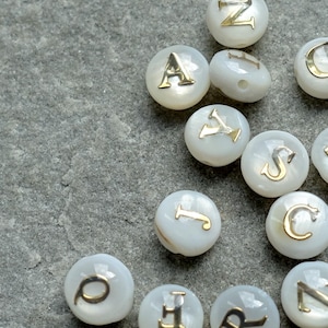 May include: White, round beads with gold-colored letters. The letters A, C, D, G, I, J, N, O, S, T, and Z are visible. The beads are scattered on a gray, textured surface.