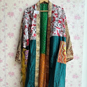 May include: A long, patchwork kimono-style robe with a variety of patterns and colors. The robe features panels of white with red floral designs, green, orange, and teal fabrics. The robe is hanging on a wooden hanger against a floral wallpaper background.